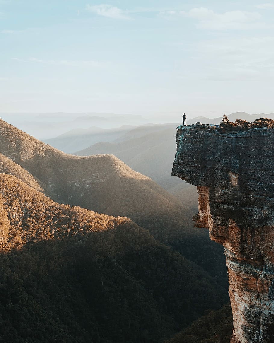 man standing on rock cliff overlooking mountains, person standing on cliff during sunrise HD wallpaper
