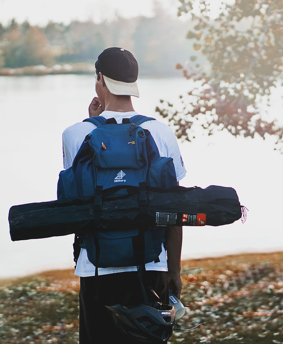 man wearing hiking backpack and tent bag standing near calm body of water, man standing near body of water selective focal photo during daytime HD wallpaper