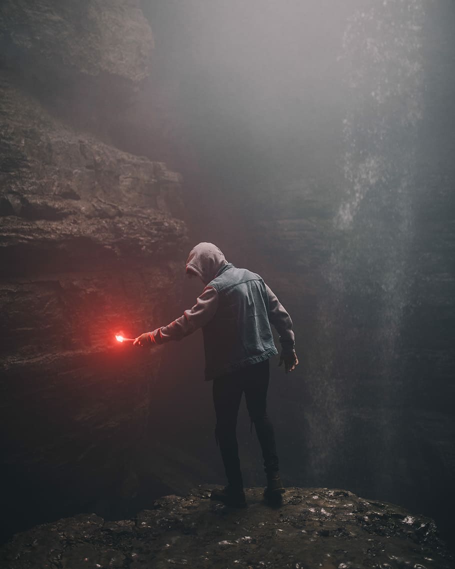 man on edge of cliff holding signal flare light, man in hooded jacket holding lighted flare in cave