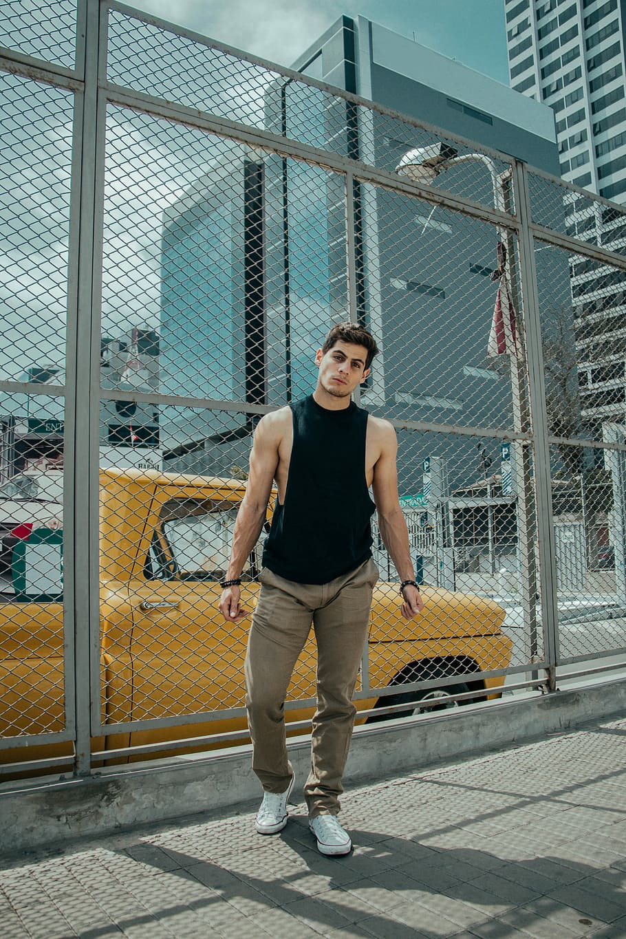 man in black tank top and brown jeans standing beside of wire fence during daytime, man standing near chain link fence distance with buildings during daytime, HD wallpaper