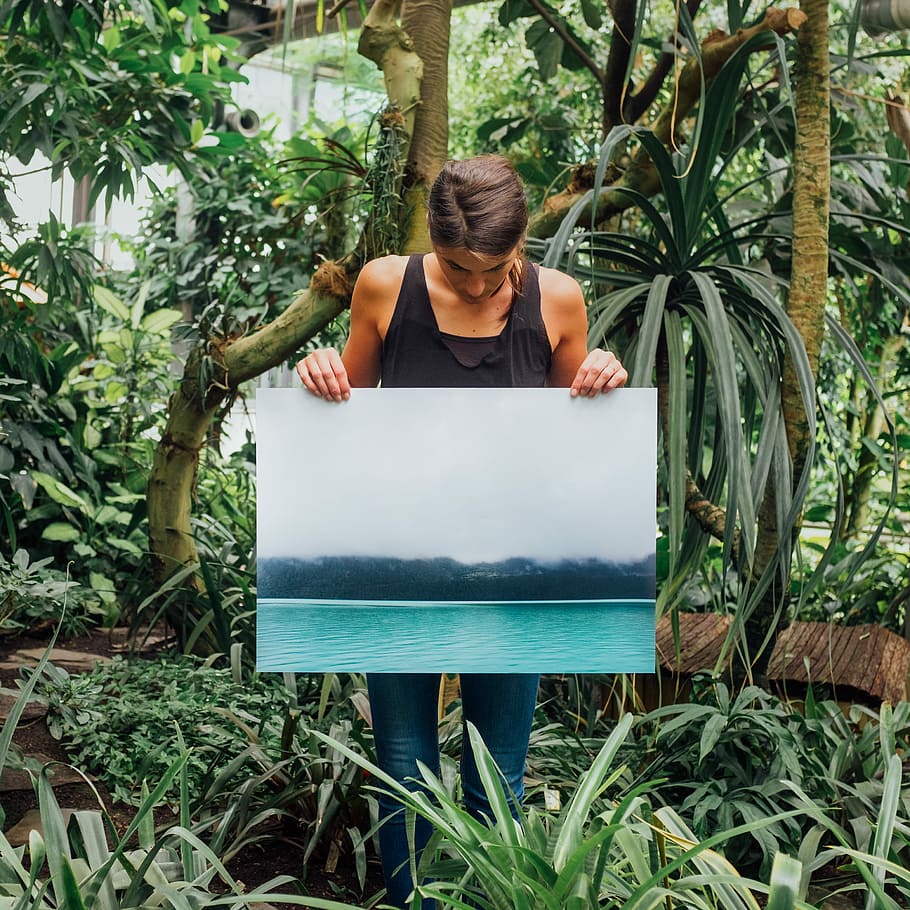 woman holding photo of tidal wave, person wearing black tank top holding poster with body of water near tree print