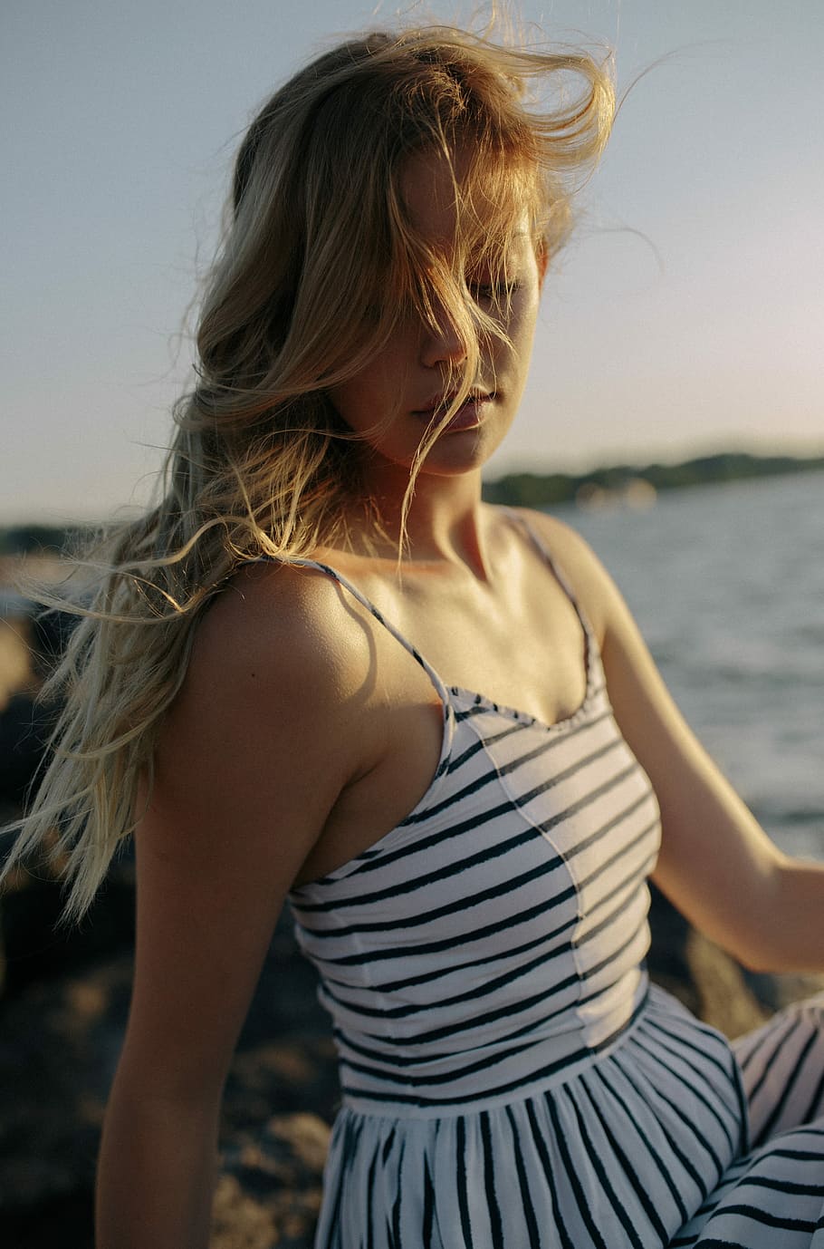 selective focus photography of woman wearing white and black striped spaghetti strap dress, shallow focus photography of a woman sitting on stone beside the beach