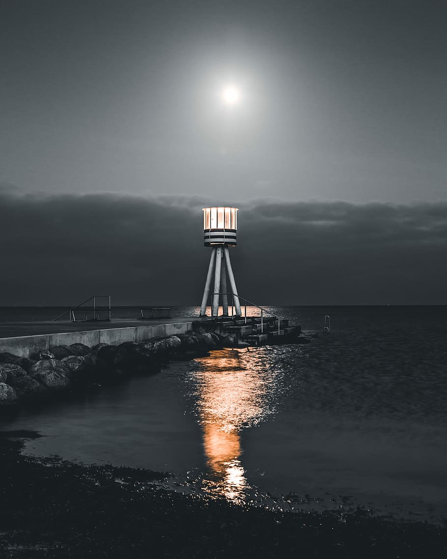 lighthouse at the dock during night, selective photo of lighthouse beside sea HD wallpaper