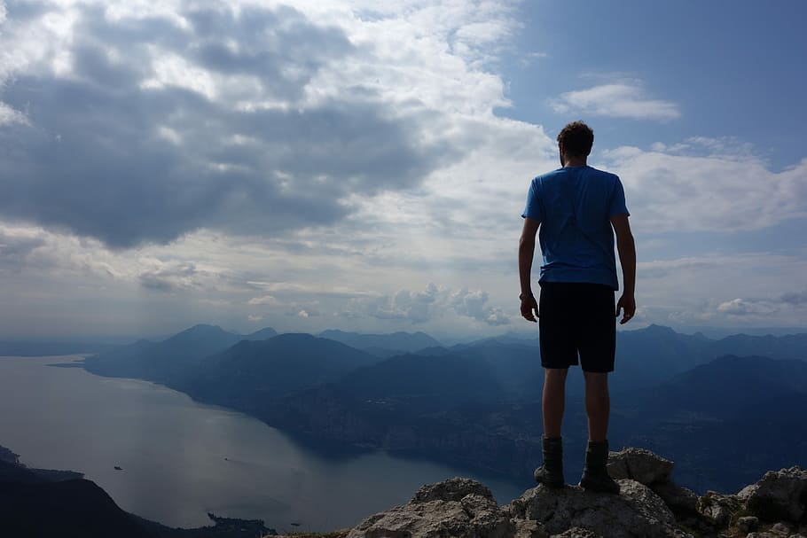 man standing on mountain cliff under cloudy sky during daytime HD wallpaper