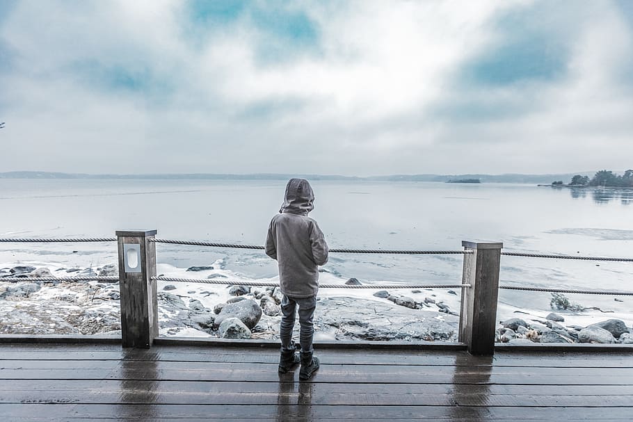 child standing infront of seashore on brown platform, person in gray hoodie standing beside of fence in front of body of water during daytime HD wallpaper
