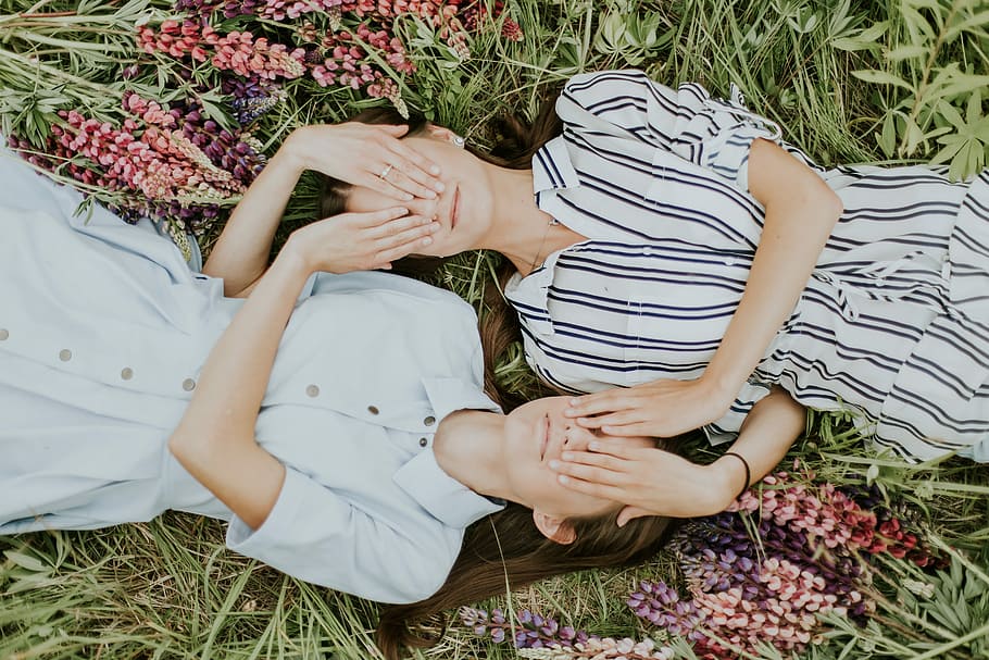 photo of women covering their eyes, two women covering each other's eye while lying on the grass