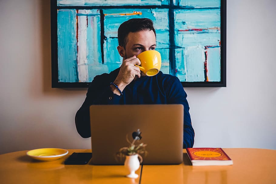 man drinking coffee in front of the laptop computer, man wearing blue dress shirt and holding yellow mug