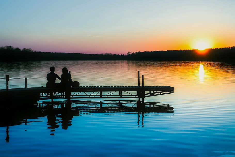 silhouette of two people sitting on docks near body of water HD wallpaper
