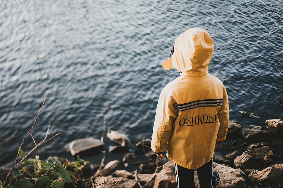 person in yellow jacket standing on cliff, boy wearing yellow hoodie facing body of water