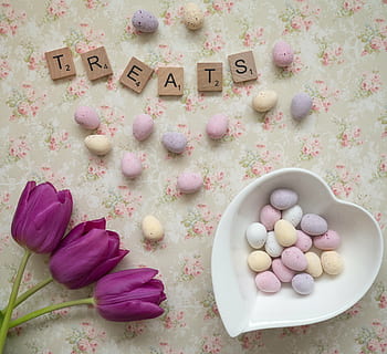 Treats scrabble letters on white, green, and pink floral surface beside chocolate eggs and heart-shaped white ceramic bowl HD wallpaper