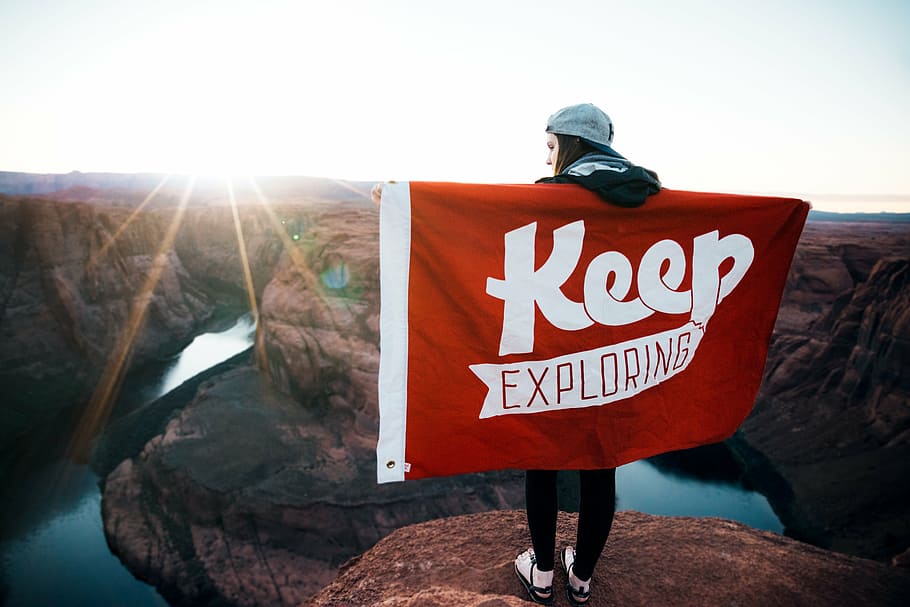 person standing on cliff holding red and white Keep exploring flag