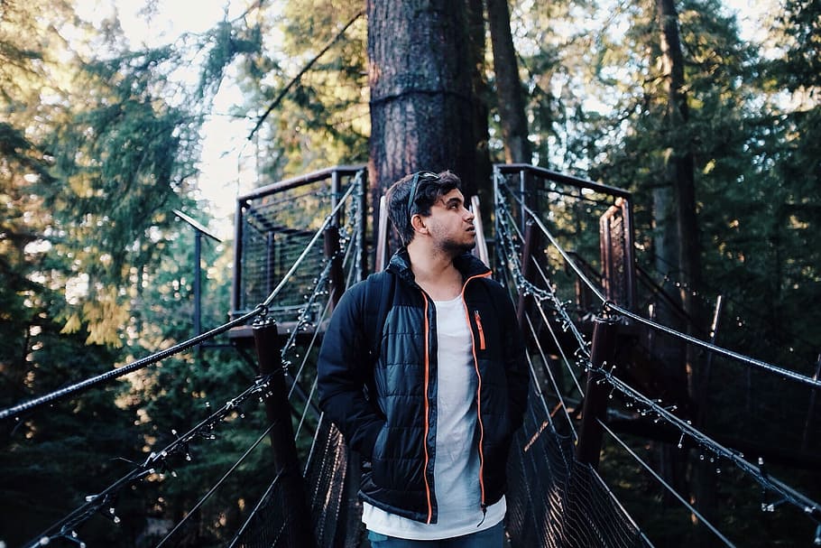 man standing on bridge in forest during daytime, man wearing black zip-up jacket standing on black hanging bridge between trees during daytime HD wallpaper