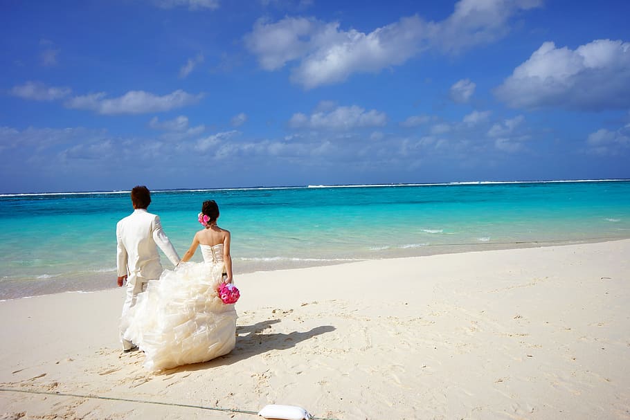 photography of couple wearing white wedding dress and suit standing on beige sand near sea under gray and blue sky during daytime