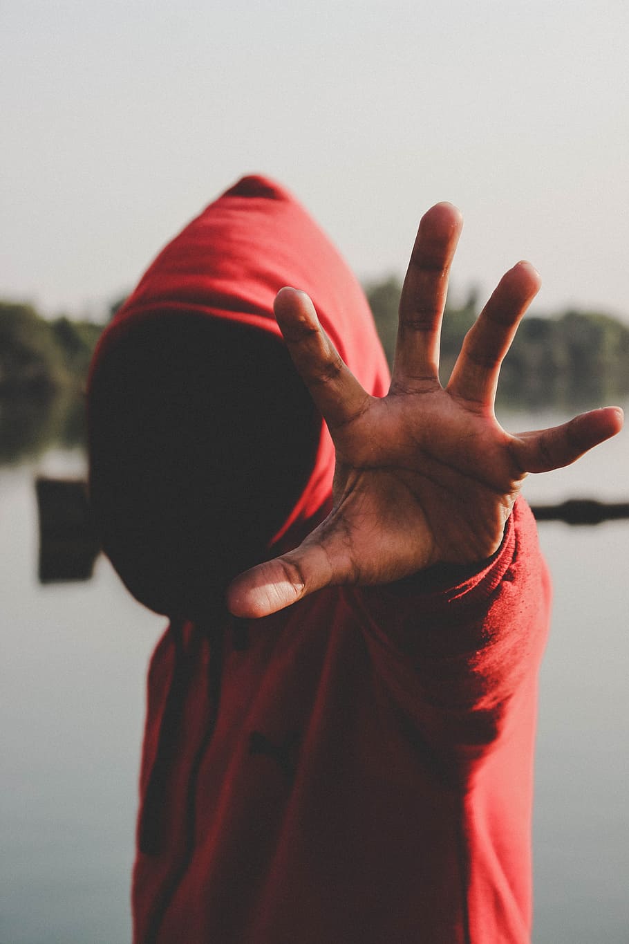 shallow focus photography of person in red hooded jacket, man wearing reaching his hand standing beside body of water