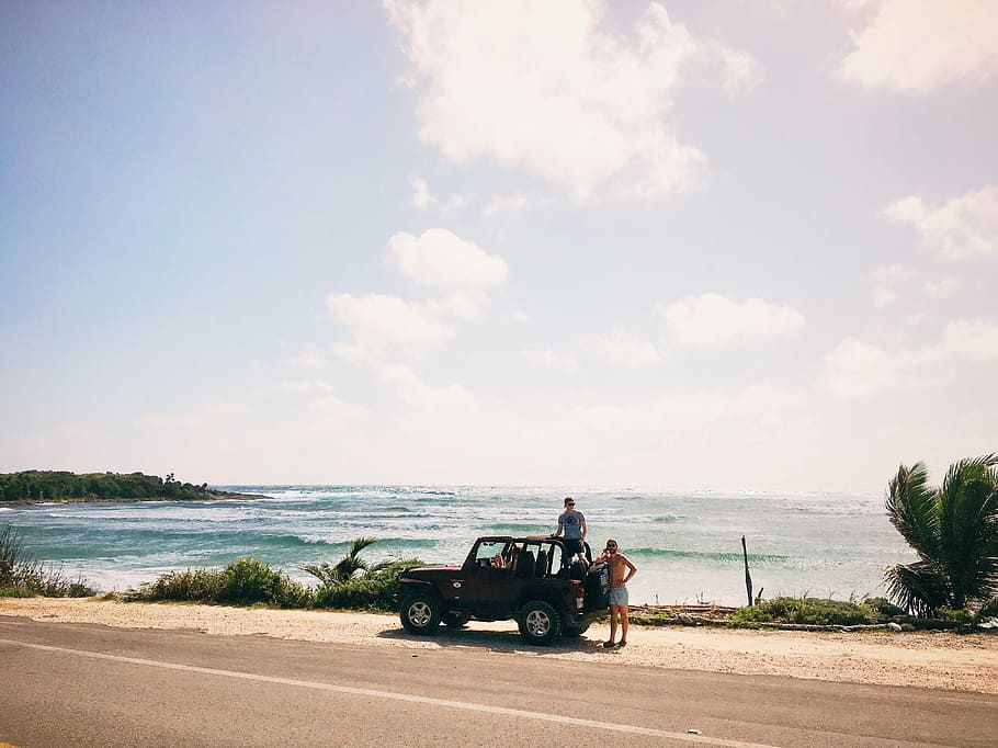 two men on brown SUV at daytime, men standing near Jeep Wrangler in a distance of sea
