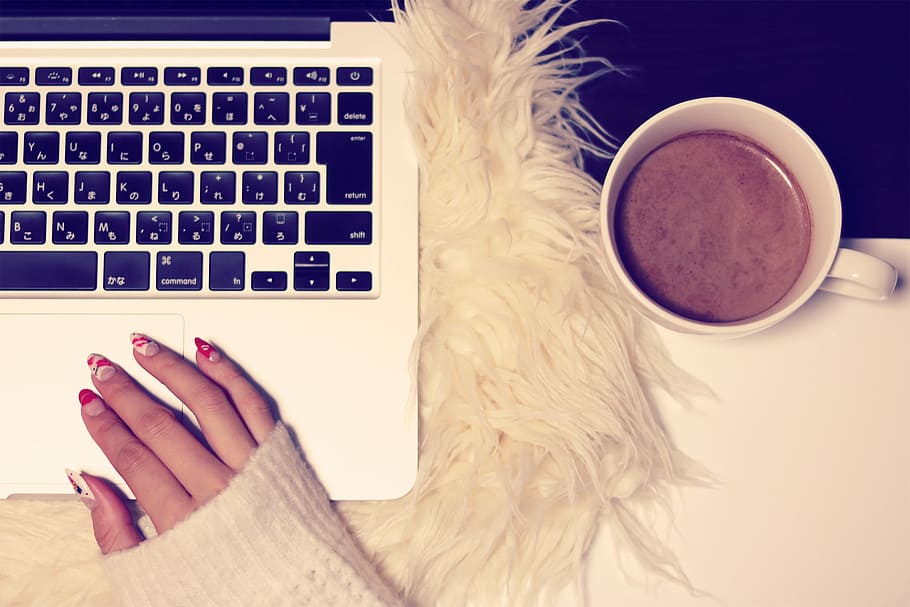 person wearing white sweater with red and white manicure on MacBook Pro beside white ceramic mug with brown liquid HD wallpaper