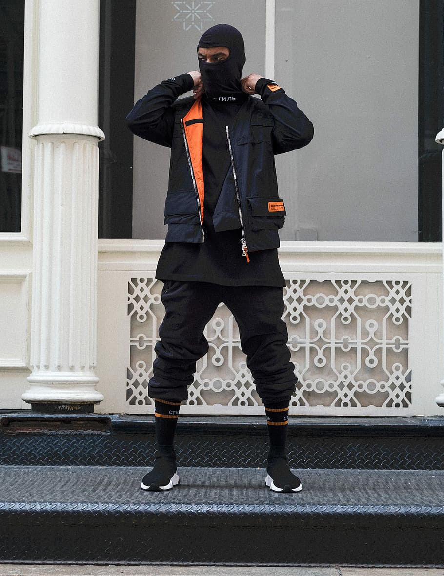 person wearing black balaclava and black and orange jacket standing near the white column