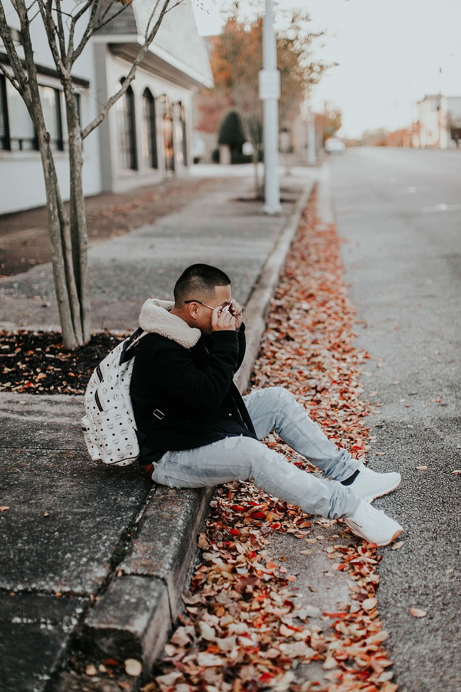 man sitting beside empty road during daytime, man in black coat wearing white backpack sitting on sidewalk during daytime HD wallpaper