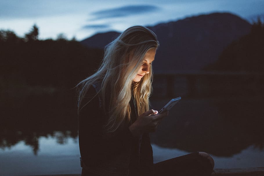 woman looking at phone beside body of water, woman in black jacket holding white smartphone