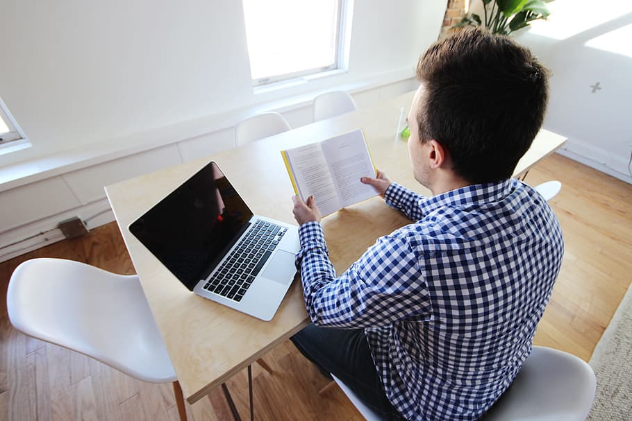 man reading book near laptop while leaning on table, macbook HD wallpaper