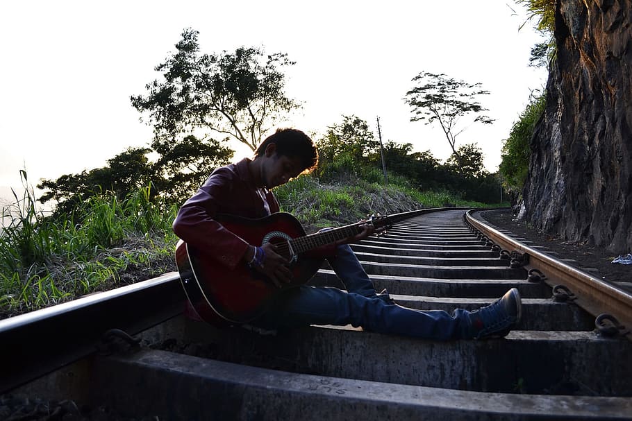 man playing a guitar sitting on train rail at daytime, railroad