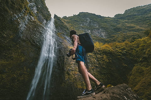 man wearing blue shorts standing on rock formation while holding DSLR camera in front of waterfalls, man looking at waterfalls HD wallpaper