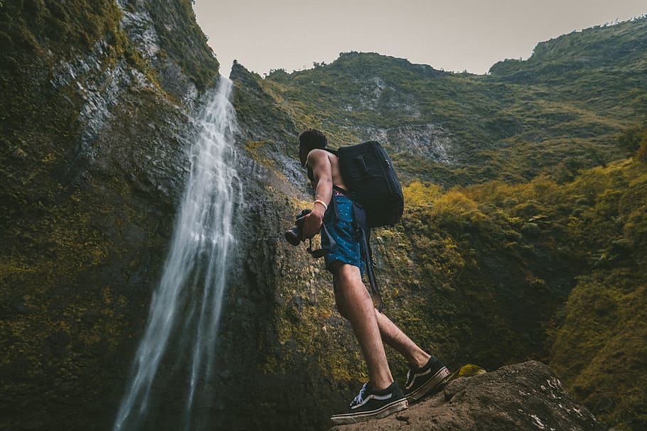 man wearing blue shorts standing on rock formation while holding DSLR camera in front of waterfalls, man looking at waterfalls HD wallpaper