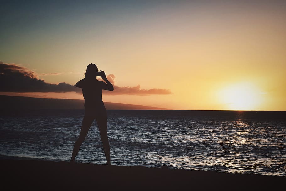 Silhouette of Woman Holding Camera Near Seashore during Golden Hour HD wallpaper