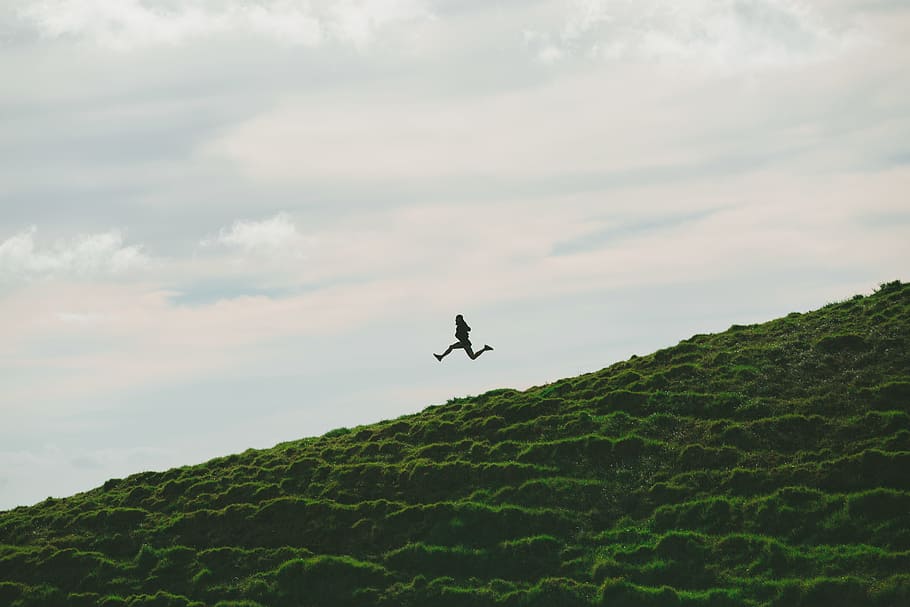 person jumping on grass field under cloudy sky during daytime, person jumping over green field