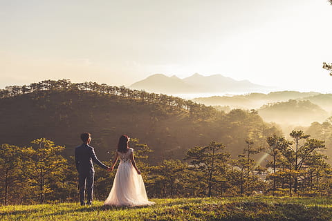 man and woman holding there hands, silhouette photo of groom and bride on green field near mountain under cloudy skies during daytime HD wallpaper