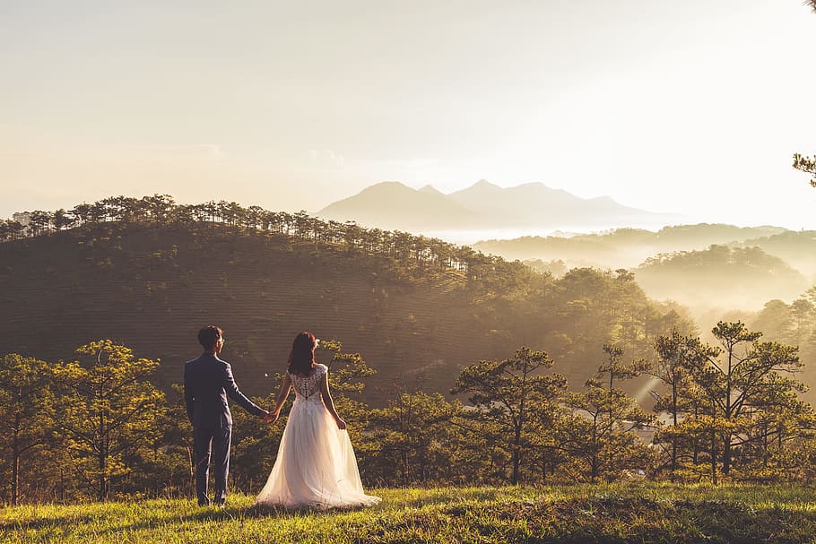 man and woman holding there hands, silhouette photo of groom and bride on green field near mountain under cloudy skies during daytime