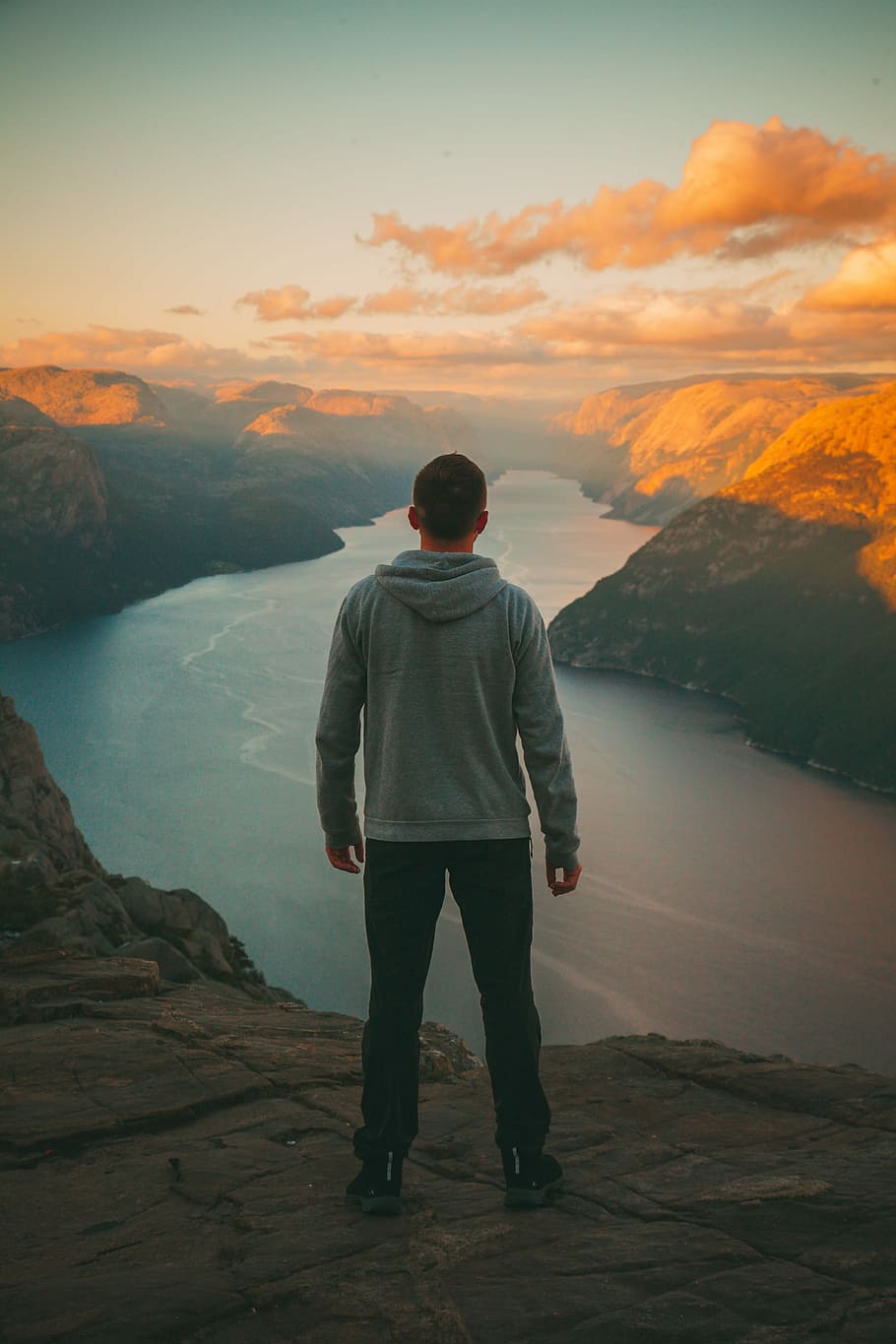 man wearing grey hoodie standing on mountain looking at river, man standing on cliff facing body of water during golden hour