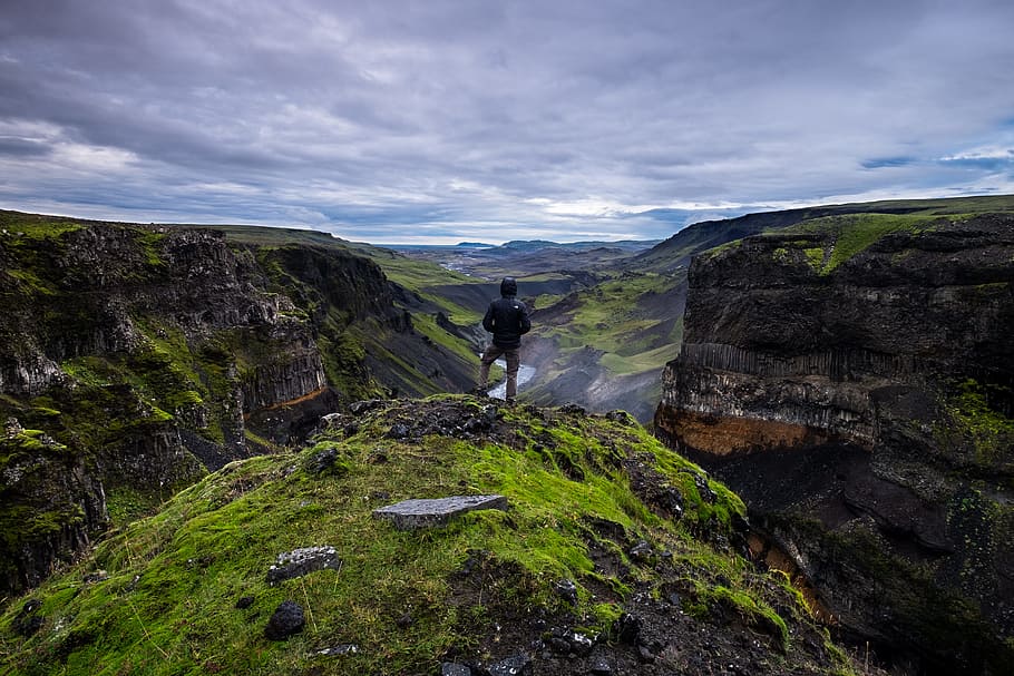 person standing on top of rocky hill viewing lake under gray clouds during daytime, person standing on cliff under gray sky at daytime HD wallpaper