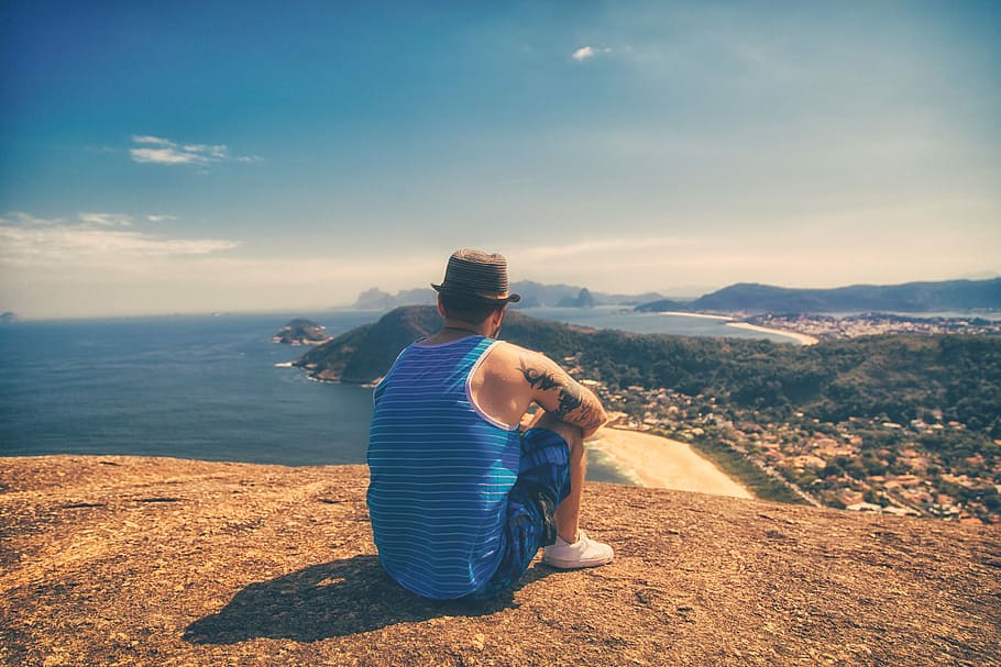 man sitting on hill facing body of water under clear sky, man sitting in mountain HD wallpaper