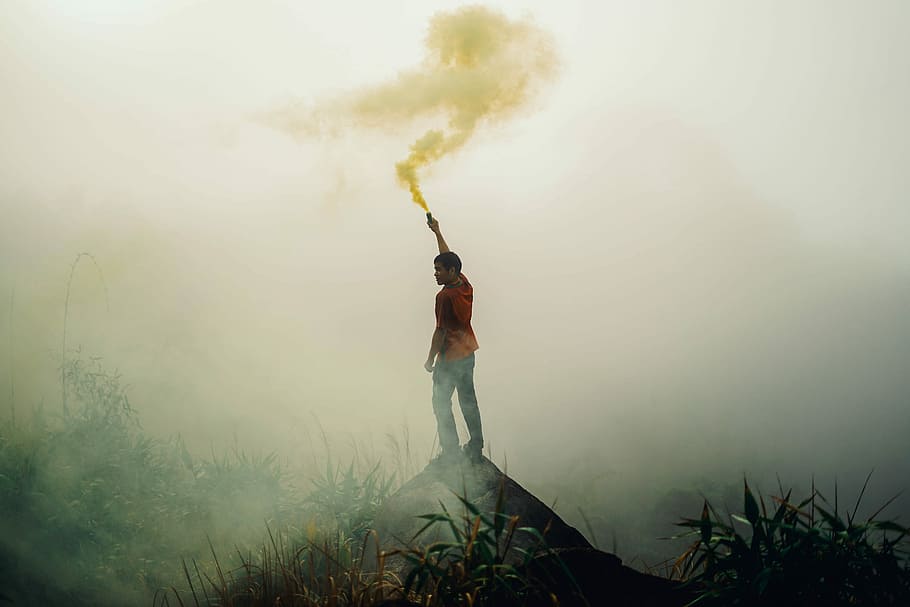 man standing on rock formation holding yellow flare surrounded by fogs, person standing on hill while holding smoke flare