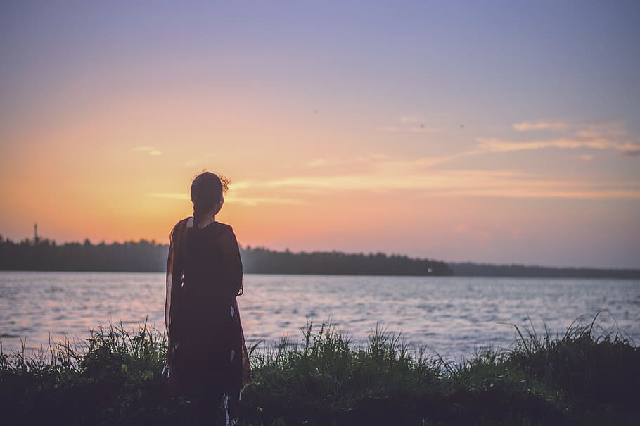silhouette photography of woman looking at body of water, woman standing alone facing sea under orange sky