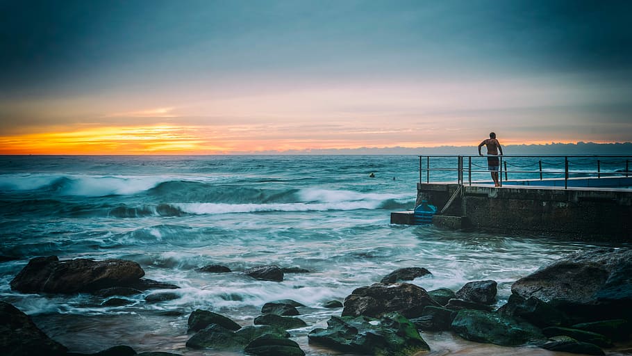 man standing on concrete dock beside seawave at daytike, sunset