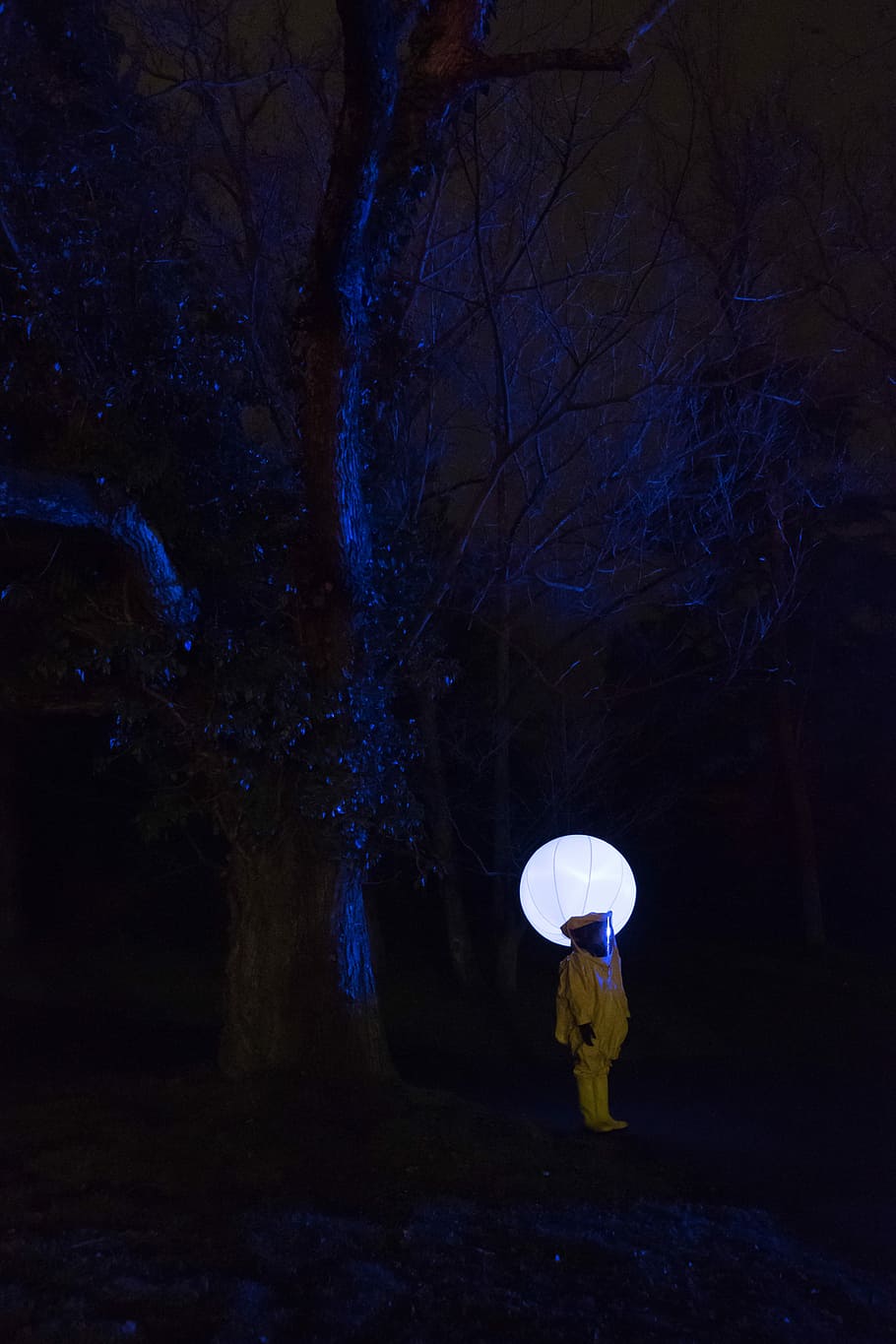 person in white outfit standing under leafless tree at nighttime, person wearing hazmat suit standing near tree