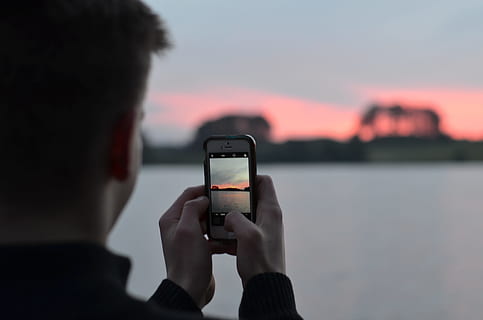 selective focus photography of person using smartphone camera taking photo of blue body of water, man holding smartphone white taking a photo of sunset beside body of water HD wallpaper