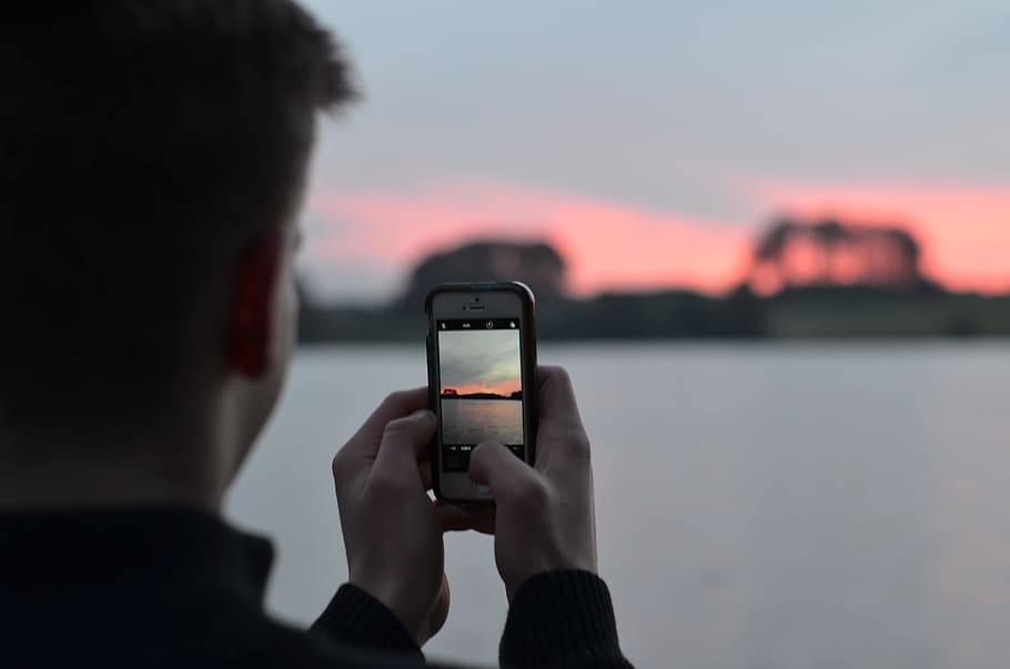 selective focus photography of person using smartphone camera taking photo of blue body of water, man holding smartphone white taking a photo of sunset beside body of water HD wallpaper