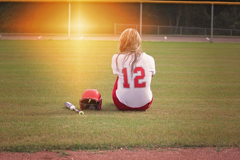 woman in white and red 12 jersey sitting on the field beside baseball bat and batting hat