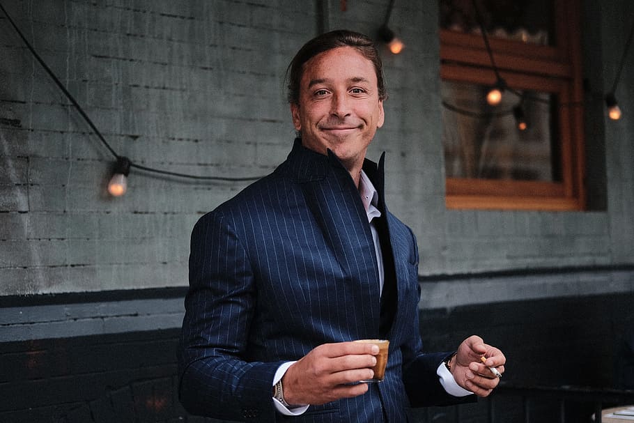 man wearing black suit holding brown mug near black string lights, man standing front of wall while smiling HD wallpaper