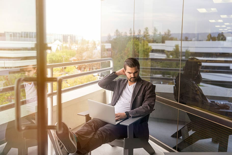 man sitting on gray arm chair using silver laptop computer on building balcony at daytime, man sitting on gray wooden Adirondack chair using gray laptop near glass wall HD wallpaper