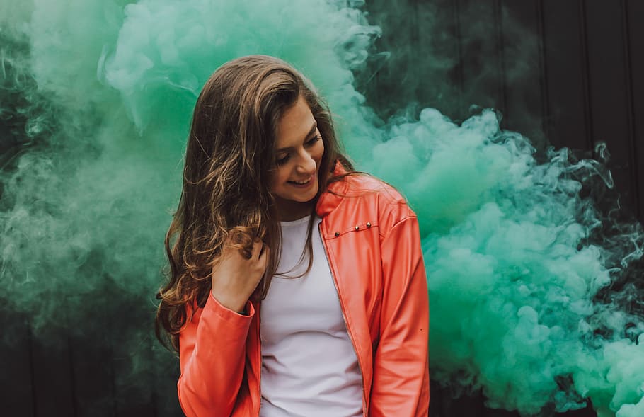woman wearing orange leather jacket surrounded by green smoke, woman smiling while holding hair standing near green smoke, HD wallpaper