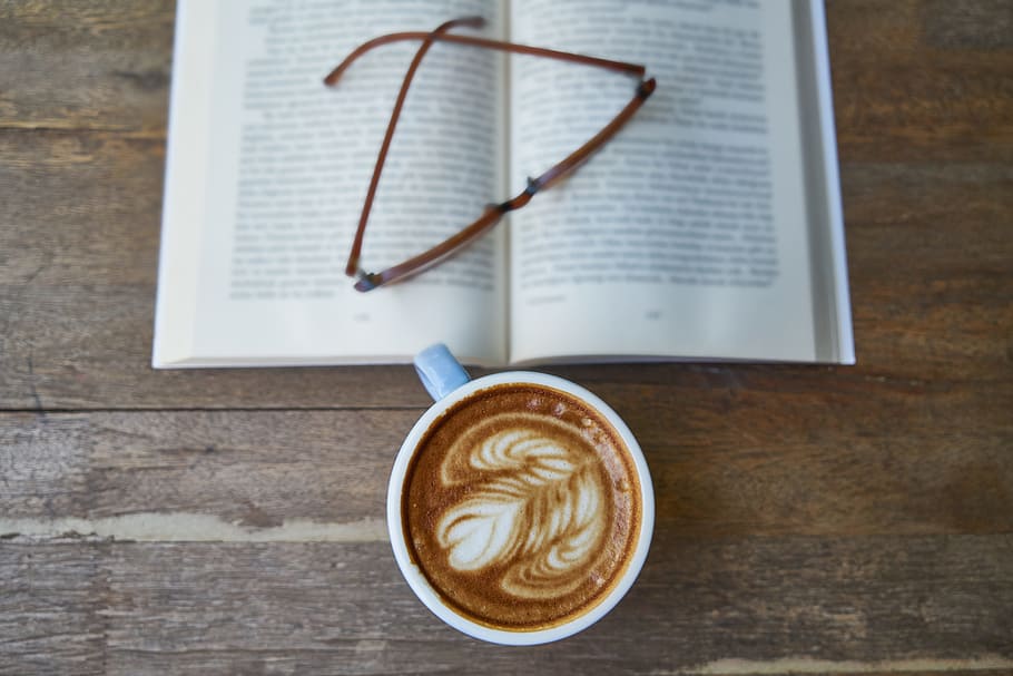 white ceramic mug filled with coffee latte beside brown framed eyeglasses on book