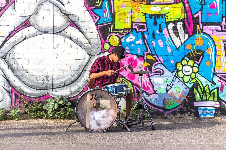 man wearing black and red checkered long-sleeved collared shirt playing drums near graffiti painted wall during daytime, HD wallpaper