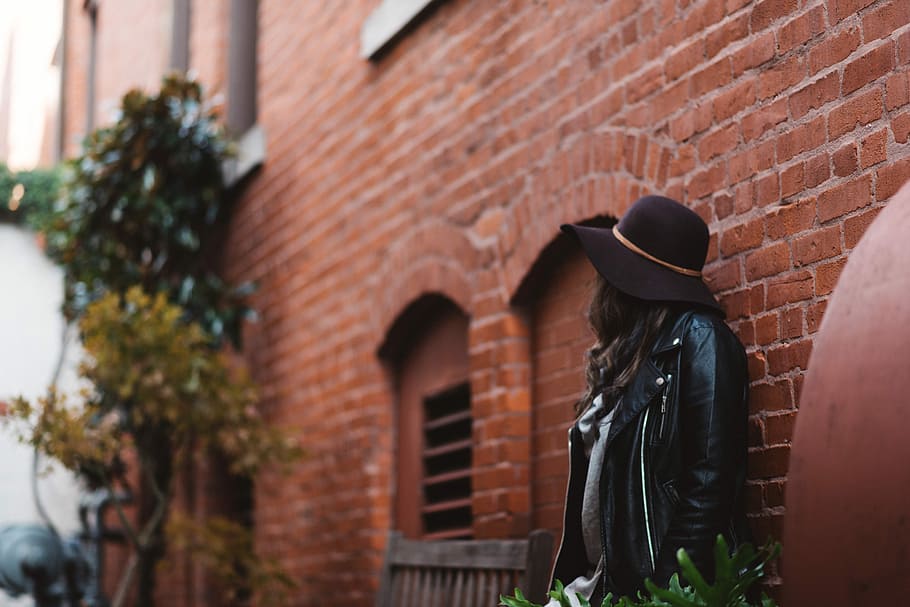 woman leaning against wall during daytime, person wearing black jacket and black cap leaning on brown brick wall HD wallpaper