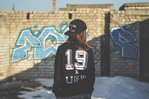 woman wearing black and white jacket and black fitted cap facing gray concrete brick wall with blue graffiti at daytime, woman standing in front of concrete blocks wall HD wallpaper