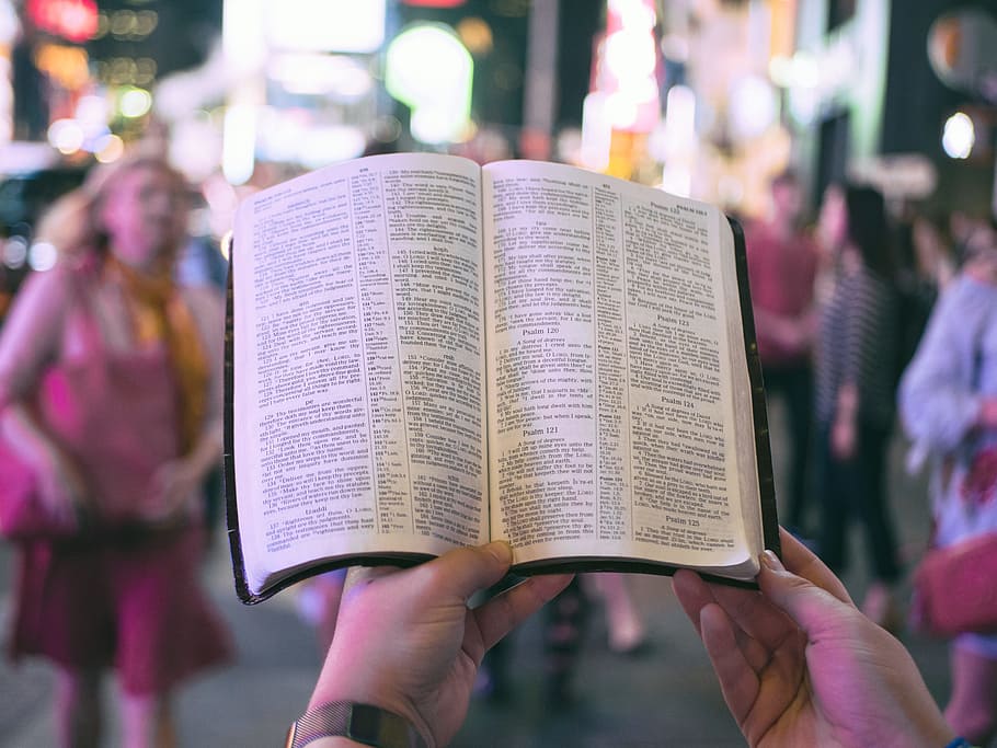 person holding bible on road with people walking on sidewalk beside buildings during nighttime, open book HD wallpaper