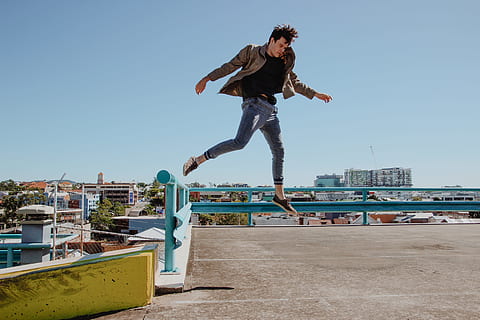 man jump from blue fence to brown concrete pavement, photography of man wearing brown jacket in open air HD wallpaper